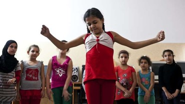 A girl dances as other children and a woman look on indoors