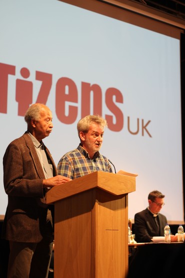 Two men speak at a lectern before a Citizens UK screen, with a clergyman seated nearby