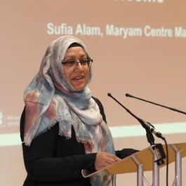 Sufia Alam speaking at a lectern, wearing a headscarf