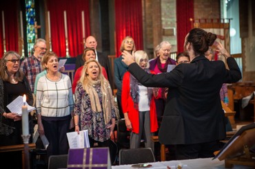 A choir sings in church, directed by a conductor, with candles held nearby
