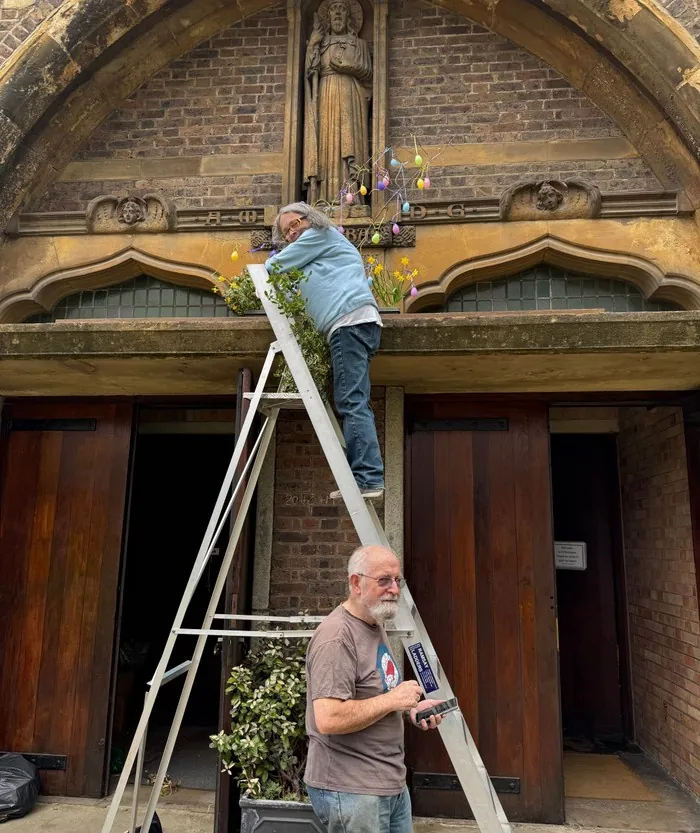 A volunteer on a ladder decorates the church porch with flowers and hanging Easter eggs