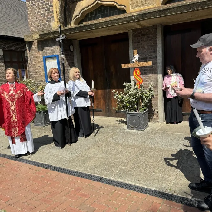 Clergy and servers in red vestments beside a wooden Pentecost cross outside the church